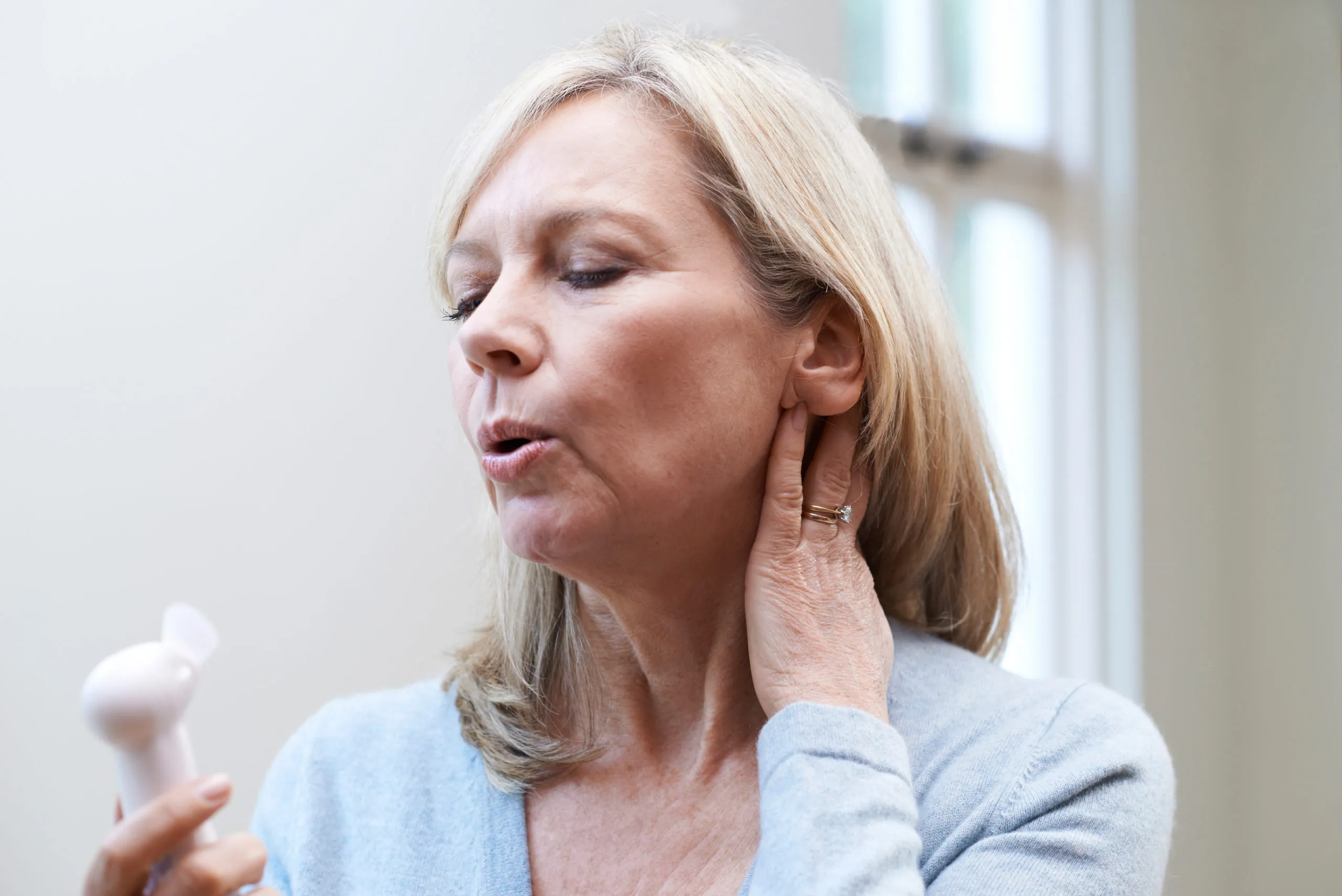 Mature woman experiencing hot flush from menopause and using a hand-held fan for relief.
