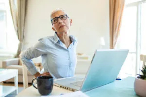 Portrait of senior stressed woman sitting at home office desk in front of laptop, touching aching back with pained expression, suffering from back pain after working on laptop