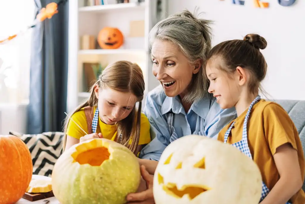 Three generations carving pumpkins for halloween, sharing laughter and creativity. Smiling girls, guided by loving grandmother, creating cherished family memories. Celebration concept