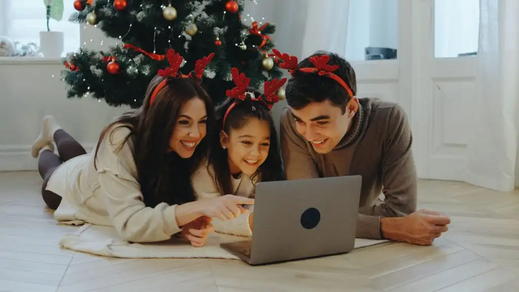 Three family members gather on the floor, wearing festive reindeer headbands, smiling and sharing a joyful moment while watching a movie on a laptop by the Christmas tree.