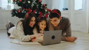 Three family members gather on the floor, wearing festive reindeer headbands, smiling and sharing a joyful moment while watching a movie on a laptop by the Christmas tree.