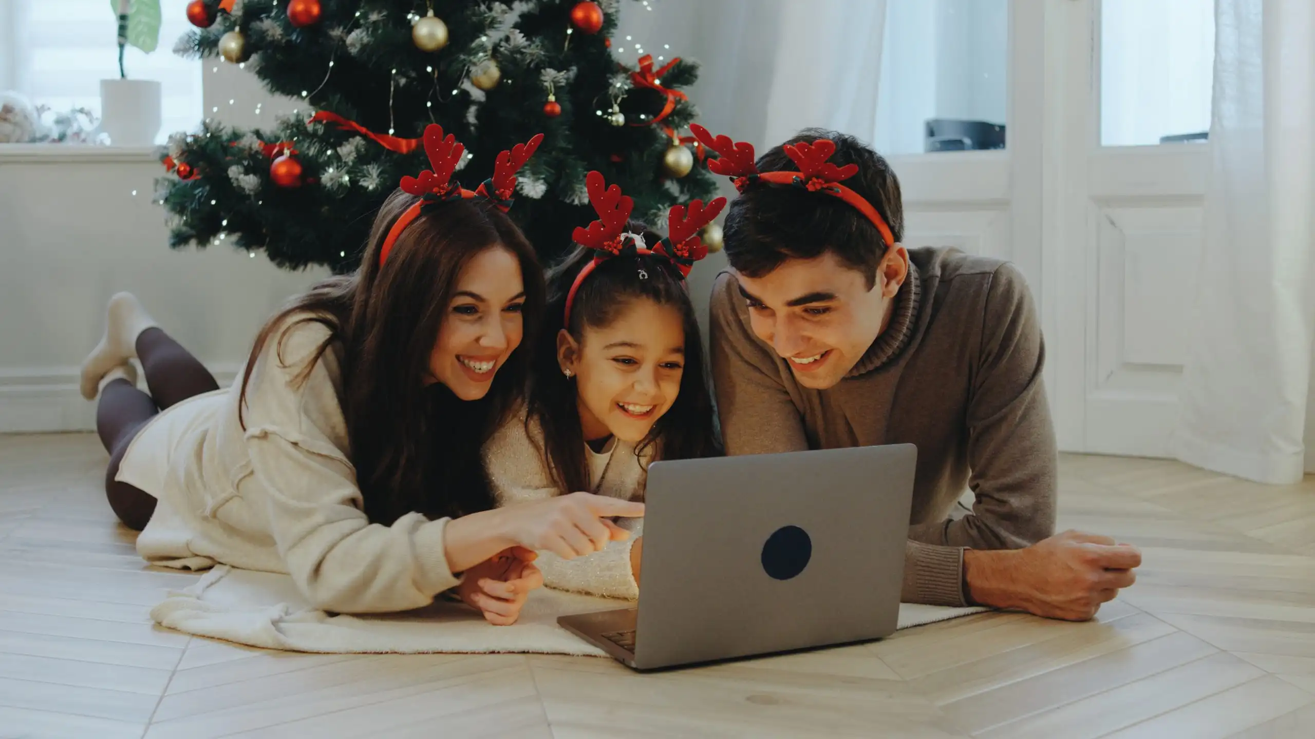 Three family members gather on the floor, wearing festive reindeer headbands, smiling and sharing a joyful moment while watching a movie on a laptop by the Christmas tree.
